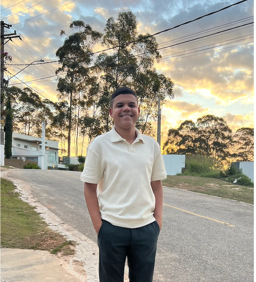Genisson Gabriel sorrindo, de camisa creme, numa rua arborizada ao pôr do sol